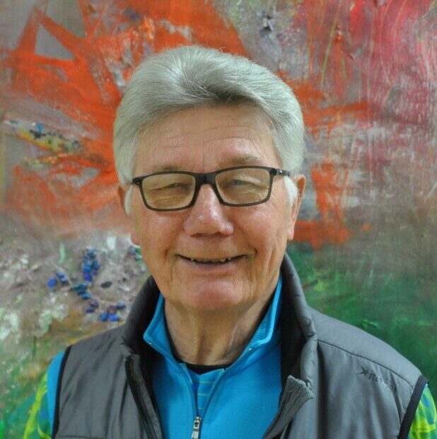 Ein lächelnder älterer Arzt. Im Hintergrund sieht man eine bemalte Wand.